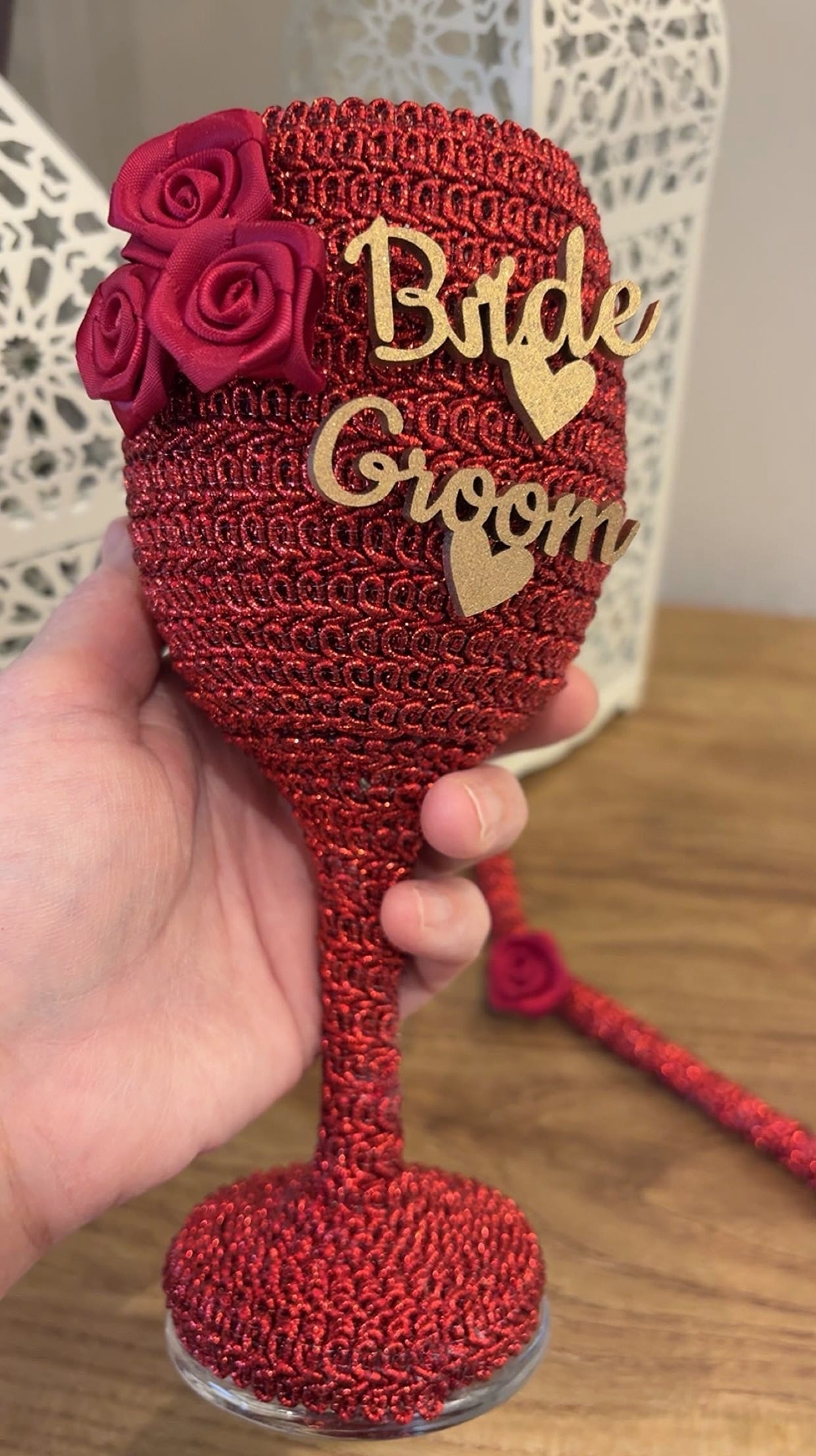 Red crochet wine glass with 'Bride' and 'Groom' text, held by a hand on a wooden surface.