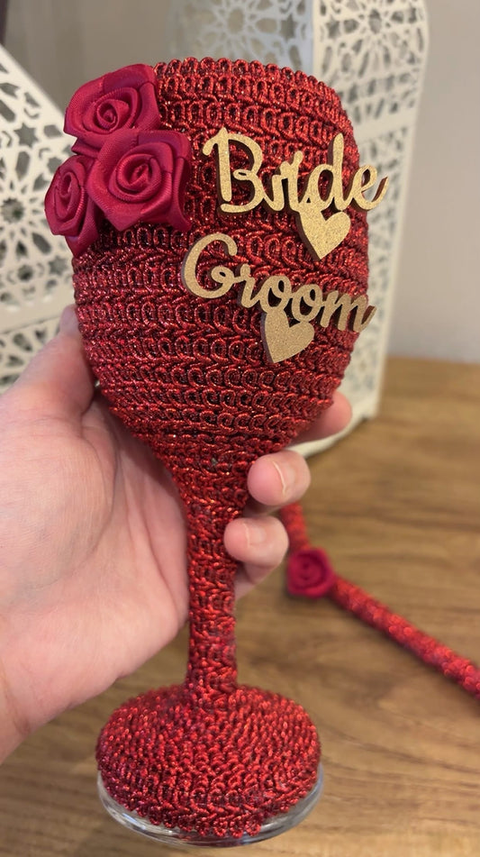 Red crochet wine glass with 'Bride' and 'Groom' text, held by a hand on a wooden surface.
